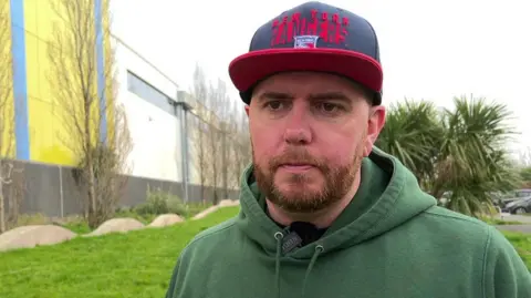 David Haddock stood outside the Plymouth Life Centre. He is wearing a green hoody and black and red hat. He has a stern look on his face as he looks slight away from the camera. 