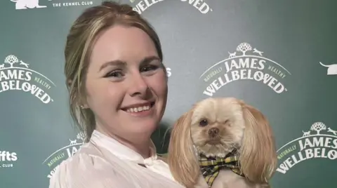 A woman smiling in a white shirt, holding a one-eye shih-tzu dog wearing a Cornish bow tie, in front of a board saying 'Scruffts'