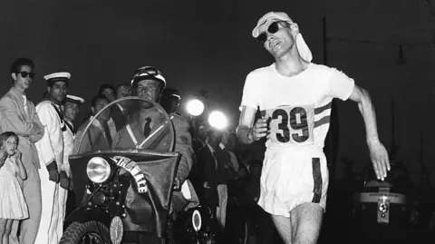 Getty Images A man in white athletics gear with the number 39 on the front, sunglasses and a white hat with a long back that covers his neck. he is closely followed by a man in a suit on a motorbike with crowds of people at the side of the road.