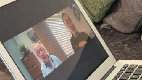 Computer screen showing 89 year old David Gompertz, alongside his daughter Hannah Gompertz. David is wearing a checked shirt and yellow heart pin badge. Hannah has chestnut brown hair tied back in a pony tail and is wearing a burgundy top with a yellow heart pin badge.