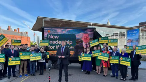 Rhun ap Iorwerth stands outside the Senedd in Cardiff Bay in front of a projector screen. He wears a black suit, pale blue shirt and a purple tie. Groups of Plaid Cymru members hold signs either side of the projector, which shows a photograph of Rhun and a sign reading 'New leadership for Wales'. Blue skies can be seen in the background.