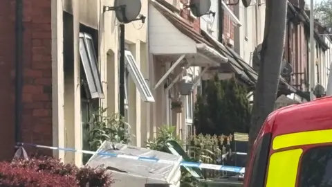PA Media A house with black smoke damage on the inside. A red fire vehicle is parked outside with blue and white police tape attached to the side of the house.