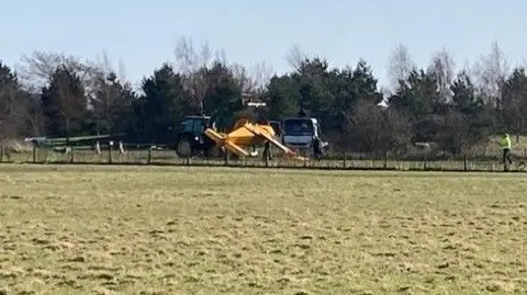 Stuart Whincup/BBC An upside down light aircraft, yellow or orange in colour, in a field. A person is stood near the crashed aircraft.