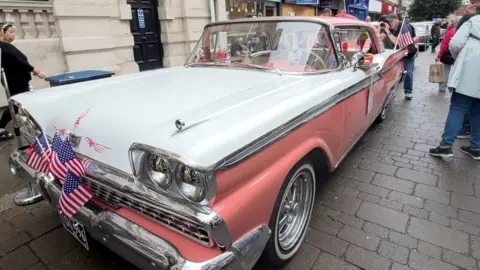 BBC A retro car on the street in Gloucester's city centre. The bodywork is a mixture of salmon pink and white and there are a handful of USA stars and stripes flags attached to the bonnet