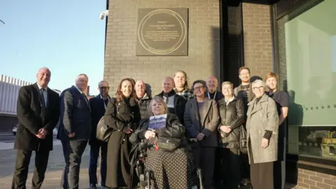 City of Wolverhampton Council A line of people are standing in front of a building. Above them is a commemorative plague. A woman in the middle of the row is in a wheelchair