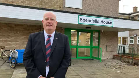 Henry Godfrey-Evans/BBC A man in a three-piece suit smiling in front of a building called "Spendells House"