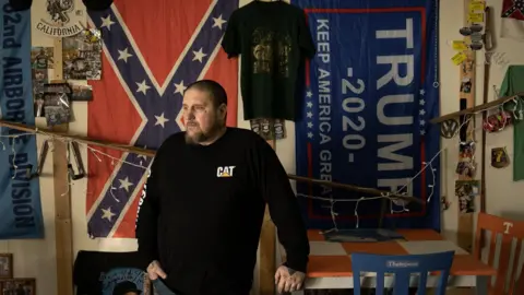 Joel Gunter/BBC Mike Macartney is pictured in front of a Confederate flag and a Trump 2020 banner. He has short hair and a beard and is leaning on a red and white checked table as he looks out of the picture