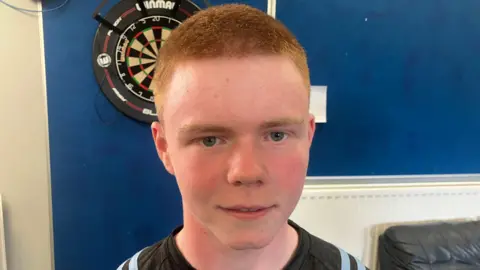 Shane O'Neill has cropped red hair and is wearing a sports top.
A dart board on the wall behind him.