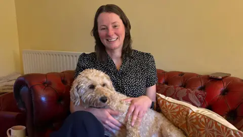 A woman with shoulder length, brown hair is smiling while sitting on a sofa. A white cockerpoo is sitting on her lap. 
