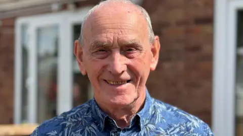 Trevor is standing in his back garden, smiling into camera. He is wearing a blue patterned shirt.