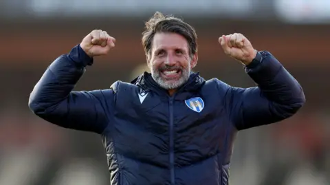PA Media Danny Cowley is wearing a navy tracksuit zip-up top, with the Colchester United crest printed on it. He is raising his arms and clenching his fists in celebration, with a big smile on his face, looking just beyond the camera lens.
