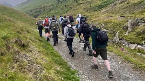 A group of men are climbing a gravel path up a hill all wearing backpacks and walking boots.