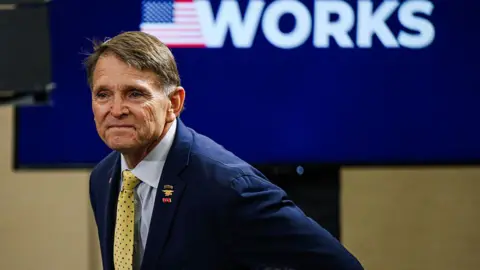 Getty Images Gallrein wears a yellow tie and suit with a sign behind him saying "America First Works"