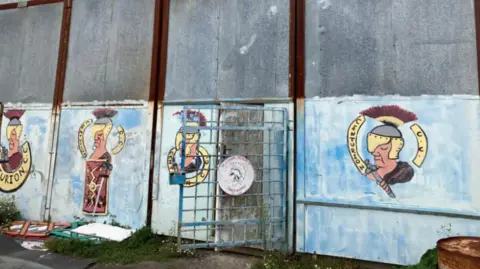 Image of the current sheds, made out of metal sheets. The bottom half of the sheets is a blue colour with centurion symbols in the middle of the blue colouring. 