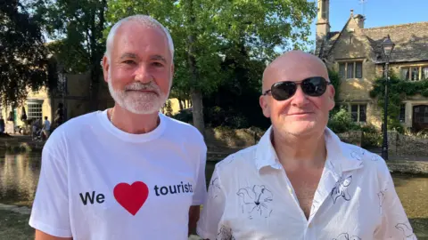 BBC Martin Steer and Paul Doxsey who are neighbours, stand next to each other. One is wearing a 'We love tourists' T-shirt and one is wearing a white shirt. They are smiling lightly at the camera. 