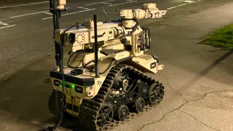 Stephen Huntley/BBC A small robot with rubber tracks, mounted cameras, coiled cables, and an articulated robotic arm positioned forward.