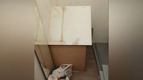 Worcestershire Regulatory Service Council officers found tobacco and other items in a hole in the store covered by a wooden box with a white lid. There is a bag of rubbish next to the box and some grey panels in front of it.
