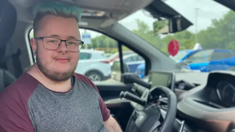 The images shows a man sat in the drivers seat of a car, looking directly at the camera. He is wearing black rimmed glasses and has a beard. He also has piercings in his nose, two on his lower lip and his right ear. He is wearing a burgundy and grey T-shirt and has blue hair.