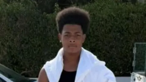 Maliki stands in front of the camera with a serious expression. He is wearing black shorts, a black vest and a white top draped across his shoulders. He appears to be on a street as cars can be seen in the background. It appears to be warm and sunny weather. 