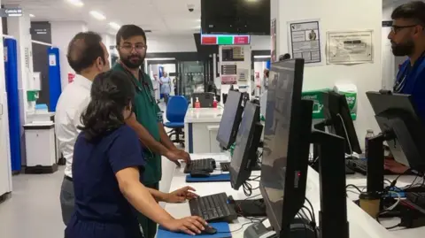 Picture shows a desk in the centre of the A&E department. Three people are stationed at computers on the left hand side of the image - one man in green scrubs, a man in a white shirt and grey trousers and a woman in blue top and trousers. At the other side of the desk, a man in blue scrubs and a stethoscope around his neck is at a computer station.