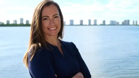Emily Gregory, the Democratic nominee for Florida state house district 87, stands in front of a waterway with buildings in the background.