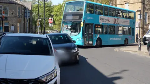 A blue bus waits on a junction behind a black car and a white car which have both stopped on double yellow lines out of frame. There is a white van waiting behind the bus too.