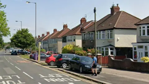 A row of semi-detached house on a dual carriageway. A number of cars are parked outside houses and a boy is about to cross the road.