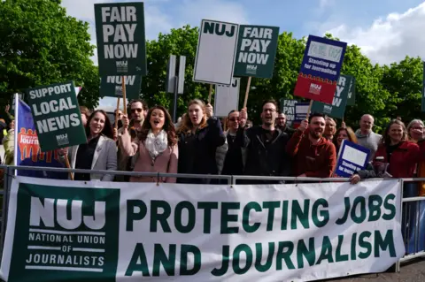 PA Media Protesters stand behind a banner reading “NUJ Protecting Jobs and Journalism” while holding placards saying “Fair Pay Now” and “Support Local News”. Members of the crowd chant and raise signs during the demonstration in sunny weather with trees in the background.