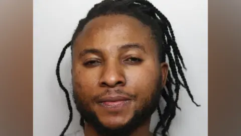 A police mugshot of Emmanuel Sakyi. He has a short black beard and his black hair is in dreadlocks. He is wearing a grey sweatshirt.