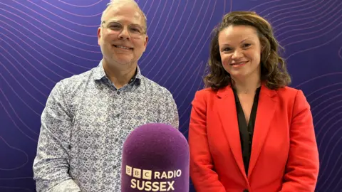 On the left presenter Danny Pike has grey hair and a grey stubble beard. He wears glasses and a patterned white and blue shirt. On the right council leader Bella Sankey has long brown wavy hair and wears a bright red suit jacket over a black top. This photo is taken in a radio studio with a purple background and a purple microphone in the foreground with the BBC Radio Sussex logo on it in white writing. 