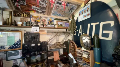 The inside of a shed with World War Two posters lining the walls, and various weaponry and memorabilia filling the space, including an old radio.