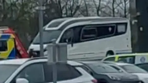 David Cardwell A white campervan in a compound. There us a white car in the foreground and other cars and police vehicles parked elsewhere.