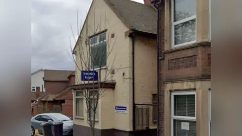 A Google image of the Greenhithe Surgery in Kent. The building is painted cream and has blue signage. It is flanked by two red-bricked houses.