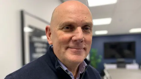 BBC Jim Gamble. He is bald wearing a navy half-zip and light blue shirt.