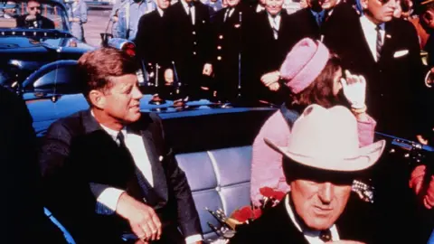 Kennedy and Mrs Kennedy in a limousine amid a crowd of people