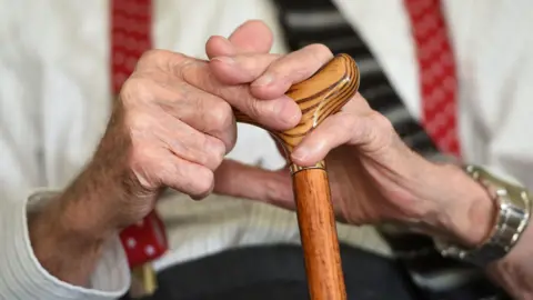 Joe Giddens/PA Wire This photograph is a close up on the hands of an elderly man holding the top of a walking stick. He is wearing a shirt, braces, tie and wrist watch