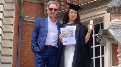 A man in a white shirt and blue suit with a woman in a degree ceremony outfit with a red brick building behind them.