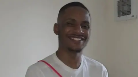 Family photo A smiling man with short dark hair and a thin moustache and beard in a white tshirt with a red diagonaly stripe in a room with white walls