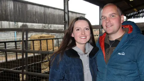 Richard and Rachel Moss, famers who are stood in a cattle shed, smile for the camera. There are hay bales and cattle pens behind them.