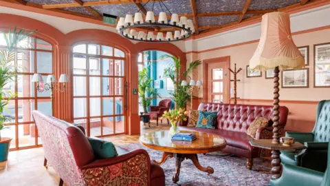 Large room with wooden windows. There are rust coloured and green sofas and chairs with flowered William Morris fabrics. There is a shiny wooden coffee table with books and Tulips on it. The walls are painted in peach and blue and there are large green plants and a chandelier along with a standard lamp and desk lamps which are wooden with cream shades. The floor is wooden with a flowered rug. The ceiling is wallpapered in blue William Morris flowered print.