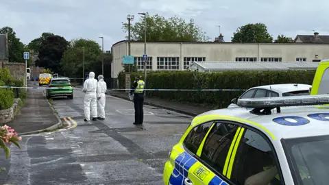 A police car is in foreground. Behind that are two people in white forensic suits, standing next to a uniformed police officer. They are behind police tape put up across a road. 
