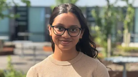 Arya is smiling at the camera. She is wearing a camel-coloured ribbed top and black-framed glasses. She has dark brown wavy hair which is tucked behind her ears and back and she has a small hoop earring in each ear. There are trees and college buildings in the background.