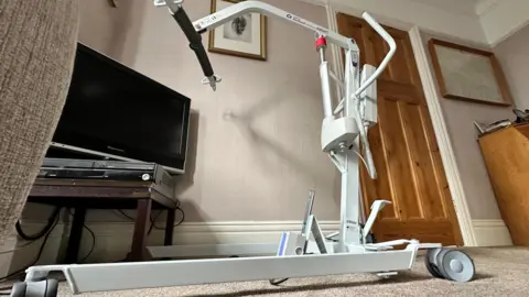 Photo of a hoist, which is a white metal machine operated by electricity and is used to lift people up so they can be moved. The hoist is sitting idle in Eric Watt's front room. 