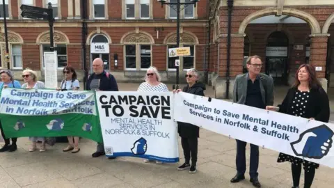 Eight campaigners from the Campaign To Save Mental Health Services in Norfolk & Suffolk stand in front of Norwich railway station holding banners