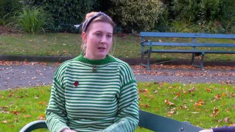 A woman with light brown hair, wearing a green and white striped jumper sits on a park bench.