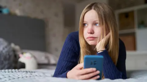 Unhappy Teenage Girl With Mobile Phone Lying On Bed At Home Anxious. She has blonde hair and is wearing a blue jumper and holding a blue phone.