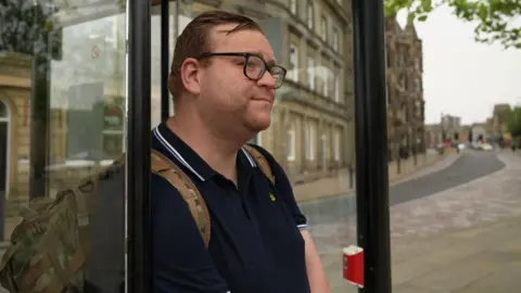 James stands at a bus stop, speaking to a reporter. He has glasses and wears a navy polo shirt.