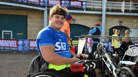 Dave Eastham is sitting in his wheelchair wearing a sports kit and trainers. He has brown hair and is wearing a blue sports top, smiling at the camera