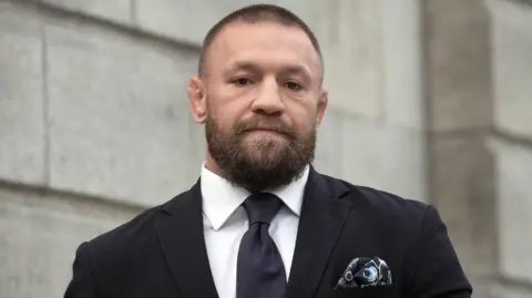 PA Media Conor McGregor is wearing a black suit with a white shirt and a black tie. He's outside a courthouse. There's a patterned pocket squire in his jacket pocket. 