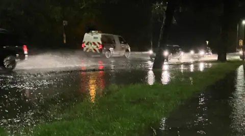 Water spray is visible near two vehicles on a road on the left of the photo in the dark. Water is also visible on a pavement on the right. 